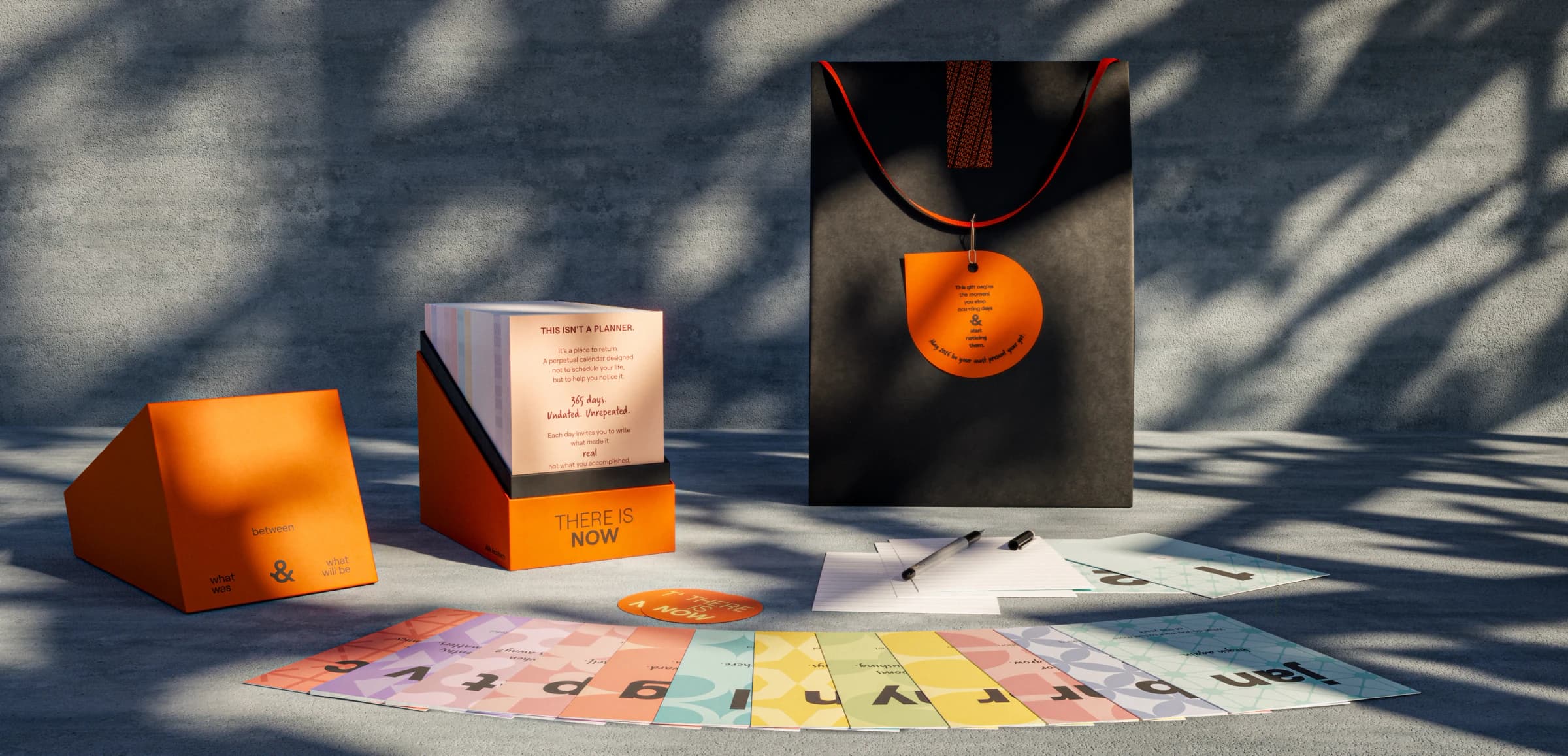 The There Is Now calendar set in sunlight: an orange box of day cards, an orange stand, a black gift bag and colorful cards
